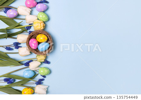 Easter colored painted eggs in bird nest with yellow daffodils, white tulips, and blue muscari on blue background with space for text and greetings. Easter and spring concept. Spring background 135754449