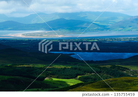 Green hills and forests meet a blue lake. Mountains fade into a bright, cloudy horizon. A vast landscape with layers of green. 135755459