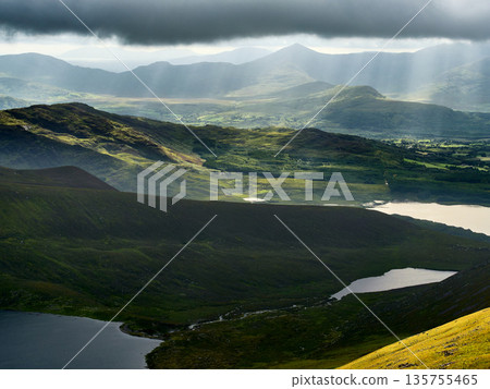 An aerial view showcases rolling green hills and several lakes under a cloudy sky in Killarney National Park. Sun rays break through, illuminating sections of the landscape. An aerial view showcases rolling green hills and several lakes under a cloudy sky in Killarney National Park. Sun rays break through, illuminating sections of the landscape. 135755465