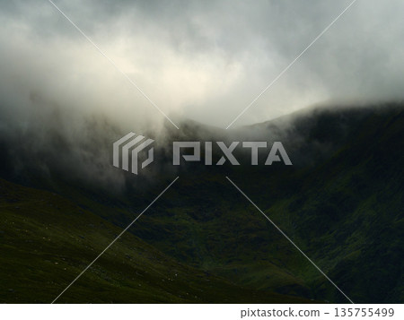 Rolling fog blankets the dark green slopes of Carrauntoohill mountain, partially obscuring the landscape. A visible walkway hints at adventure through the dramatic, misty Irish scenery. Rolling fog blankets the dark green slopes of Carrauntoohill mountain, partially obscuring the landscape. A visible walkway hints at adventure through the dramatic, misty Irish scenery. 135755499