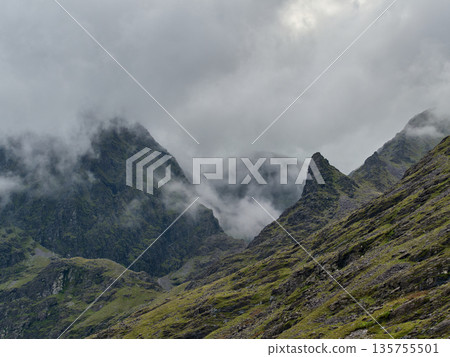 Majestic mountains rise through thick mist, their rugged peaks partially hidden by low clouds. Steep, green-tinted rocky slopes dominate the foreground on an overcast day. 135755501