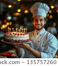 Smiling male pastry chef white uniform presents a cherry-topped birthday cake with lit candles in a warmly lit kitchen. Elegant and appetizing composition. 135757260