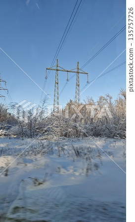 Electric power transmission towers standing above snowy bushes and trees under a clear blue winter sky. Image represents energy infrastructure, cold season landscape and connection between technology 135757726