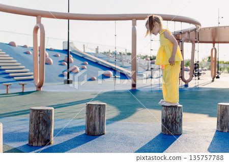 Little girl outfit balancing on playground stumps, focused and active on sunny day. 135757788