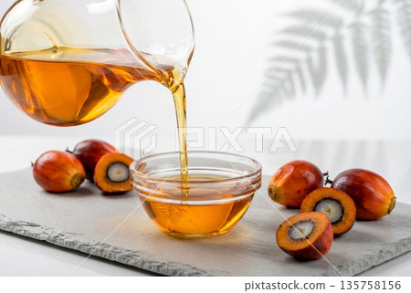 Pouring golden palm oil from glass pitcher into bowl with fresh red palm fruits on slate 135758156