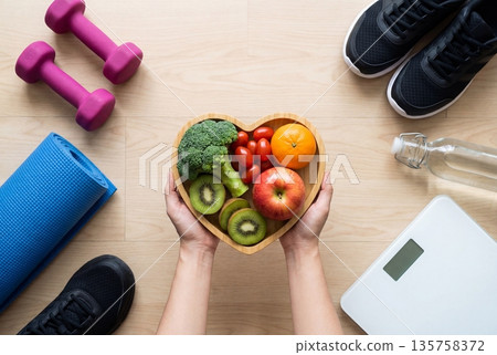 Hands holding heart shaped bowl of healthy food near dumbbells sneakers and yoga mat on wooden floor 135758372