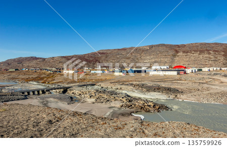 Inland arctic greenlandic settlement with river, bridge and scattered buildings surrounded by rocky terrain and dry tundra landscape in Kangerlussuaq, Greenland 135759926
