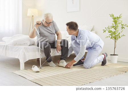 Young nurse or health care worker helping a senior patient with crutches to put on shoes at home. Young nurse or health care worker helping a senior patient with crutches to put on shoes at home. 135763726