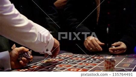 Gambling Game Scene, Individuals Strategically Placing Chips Around Roulette Table On Lowlit Gambling Floor 135764491