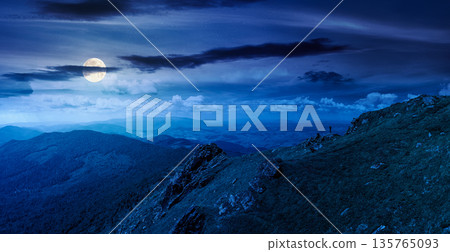 breathtaking summer mountain landscape at night. rocks on edge of hill in full moon light. view in to valley. grassy slope under cloudy sky. distant ridge on the horizon. epic mysterious fantasy 135765093
