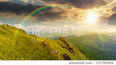 breathtaking summer mountain landscape at sunset. rocks on edge of hill in evening light. view in to valley. grassy slope under cloudy sky. distant ridge on the horizon. epic weather and rainbow 135765099