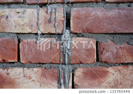 Untidy brickwork with gaps between bricks unfilled with cement mortar. Gray cement stains like melted candle wax on the hand-molded red brick. Close-up. Abstract background in the style of punk rock. 135765449