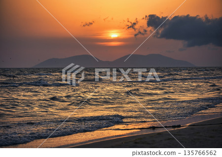 Vlora, Albania  August , 2025 A serene sunset over the Adriatic Sea, with warm golden light reflecting across gentle waves and distant silhouettes of mountains. The dramatic clouds and soft evenin 135766562