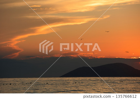 Vlora, Albania  August , 2025 A serene sunset over the Adriatic Sea, with warm golden light reflecting across gentle waves and distant silhouettes of mountains. The dramatic clouds and soft evenin 135766612