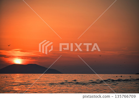 Vlora, Albania August , 2025 A serene sunset over the Adriatic Sea, with warm golden light reflecting across gentle waves and distant silhouettes of mountains. The dramatic clouds and soft evenin 135766709
