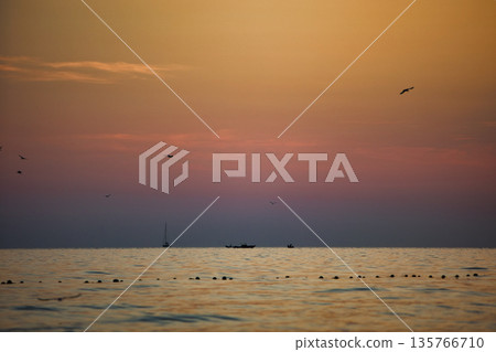 Vlora, Albania August , 2025 A serene sunset over the Adriatic Sea, with warm golden light reflecting across gentle waves and distant silhouettes of mountains. The dramatic clouds and soft evenin 135766710