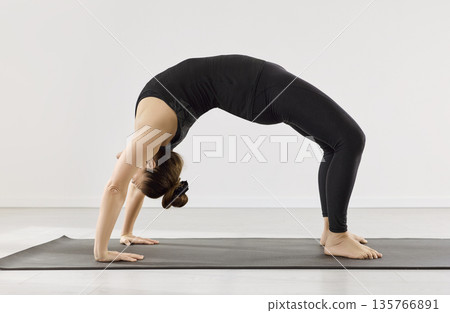 Woman practicing wheel pose on yoga mat in minimal studio, side view indoors Woman practicing wheel pose on yoga mat in minimal studio, side view indoors 135766891