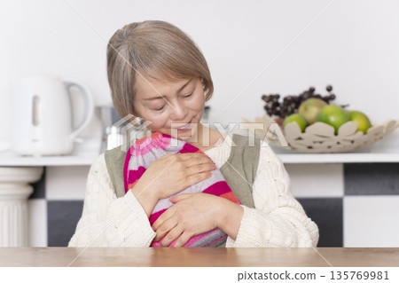 Senior woman pouring hot water into a hot water bottle in the kitchen Senior woman pouring hot water into a hot water bottle in the kitchen 135769981