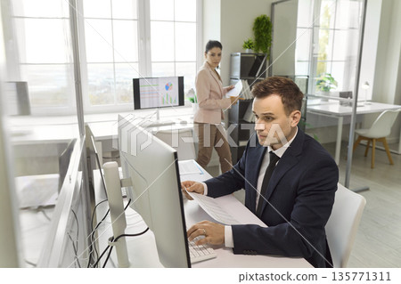 Focused businessman at desktop in modern office, analyzing data and charts Focused businessman at desktop in modern office, analyzing data and charts 135771311
