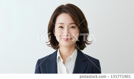 A woman in her 50s wearing a navy jacket smiling gently against a white background 135771901