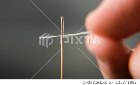 Macro shot of a hand threading white thread through an eye of needle. Symbol of precision, patience, and handmade craft concept  135771982