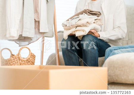 A man moving and tidying up his living room, putting his clothes into cardboard boxes as part of a decluttering project 135772163