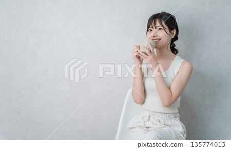 A young Asian woman in a tank top drinking a hot drink from a mug to keep warm 135774013
