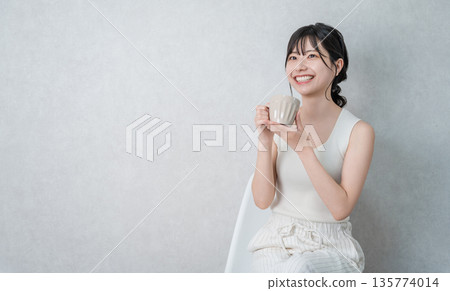 A young Asian woman in a tank top drinking a hot drink from a mug to keep warm 135774014