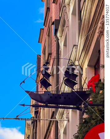Sign of a ship hangs from a building in a city under a blue sky Sign of a ship hangs from a building in a city under a blue sky 135774017