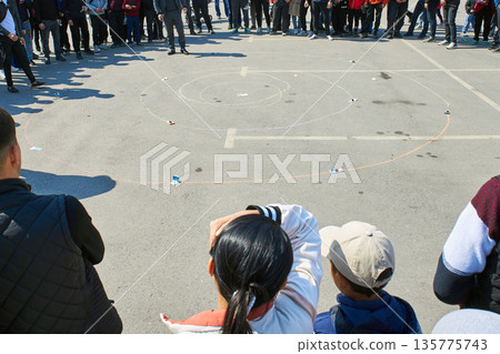 A group of people watching an ethnic game on asphalt during a Nooruz festival. Traditional nomadic game of Alchiki played with knuckles. Bishkek, Kyrgyzstan. A group of people watching an ethnic game on asphalt during a Nooruz festival. Traditional nomadic game of Alchiki played with knuckles. Bishkek, Kyrgyzstan. 135775743