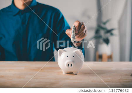 Global economy money bank finance concept helps people save and invest in stock for growth while man puts cash into white piggy bank on wooden table for business economic plans Global economy money bank finance concept helps people save and invest in stock for growth while man puts cash into white piggy bank on wooden table for business economic plans 135776682