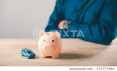 Man planning car money bank insurance piggy save auto loan cost finance fuel saving tax, sitting with arms crossed behind pink piggy bank and blue toy vehicle on light wooden table top Man planning car money bank insurance piggy save auto loan cost finance fuel saving tax, sitting with arms crossed behind pink piggy bank and blue toy vehicle on light wooden table top 135777464