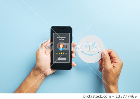 Review online customer feedback, hand holds smartphone displaying five star rating for positive service experience, another hand holds smiling face indicating high satisfaction 135777469