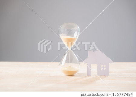 Hourglass showing passing time next to white house cutout represents home mortgage loan concept, illustrating property buying investing sell building accommodation temporary solution for estate 135777484