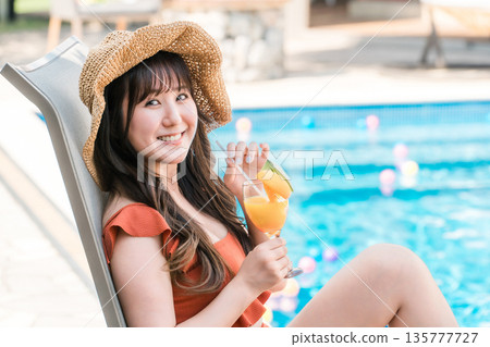 一位年輕的亞洲女子穿著泳裝，坐在泳池邊的躺椅上喝著熱帶果汁。 135777727