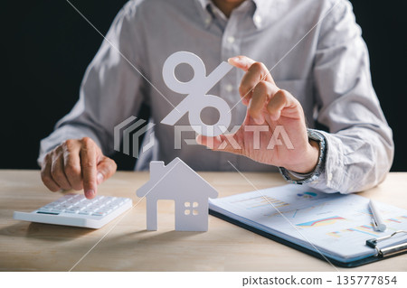 Businessman calculates home property percent rate, holding large percentage symbol above small house model, mortgage loan charts and financial data, planning real estate investment interest tax Businessman calculates home property percent rate, holding large percentage symbol above small house model, mortgage loan charts and financial data, planning real estate investment interest tax 135777854