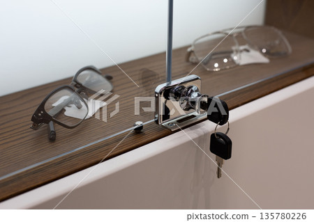 Glass cabinet with glasses behind closed transparent doors. Lock and key on display cabinet door. Closed cabinet with glasses. Optical store with shelves of glasses for visually impaired 135780226