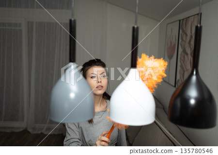 Woman Using Duster to Clean Hanging Lamps in a Modern Apartment Woman Using Duster to Clean Hanging Lamps in a Modern Apartment 135780546