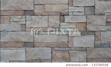 A wall finished with stone tiles in squares of various sizes in gray-brown colors with a textured surface of natural stone relief, a decorative material for finishing interior and exterior surfaces A wall finished with stone tiles in squares of various sizes in gray-brown colors with a textured surface of natural stone relief, a decorative material for finishing interior and exterior surfaces 135780598