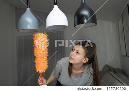 Woman Using a Feather Duster Cleaning Hanging Lamps in an Apartment Woman Using a Feather Duster Cleaning Hanging Lamps in an Apartment 135780600