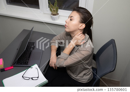 Young Woman Feeling Neck Discomfort While Working at a Desk in a Home Office During the Evening 135780601