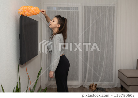Woman Dusting Television in Living Room With Dog Nearby in a House During the Day Woman Dusting Television in Living Room With Dog Nearby in a House During the Day 135780602