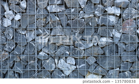 A fragment of a gabion with a metal cage filled with large gray boulders, the hard, solid texture of stone and metal, a view of a finished gabion with natural rock filling behind an iron grate A fragment of a gabion with a metal cage filled with large gray boulders, the hard, solid texture of stone and metal, a view of a finished gabion with natural rock filling behind an iron grate 135780838