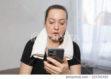 Woman Checking Phone While Wearing a Towel Around Her Neck at Home After Workout Session 135781497