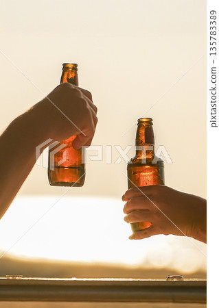 Two bottles of beer in his hands on the beach against the sunset. Friends clink glasses and relax watching the sunset, close-up. High quality photo 135783389
