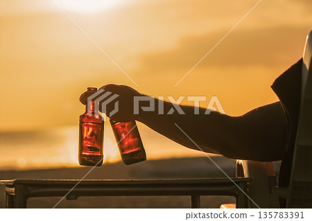 A man holds two bottles of beer in his hand at sunset on the beach by the sea silhouette. The party will start soon. He watches the sunset and enjoys his vacation. High quality photo A man holds two bottles of beer in his hand at sunset on the beach by the sea silhouette. The party will start soon. He watches the sunset and enjoys his vacation. High quality photo 135783391