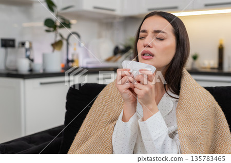 Young woman wrapped in blanket sneezes uses tissue cold flu symptoms sniffles unwell sick tired weak Young woman wrapped in blanket sneezes uses tissue cold flu symptoms sniffles unwell sick tired weak 135783465