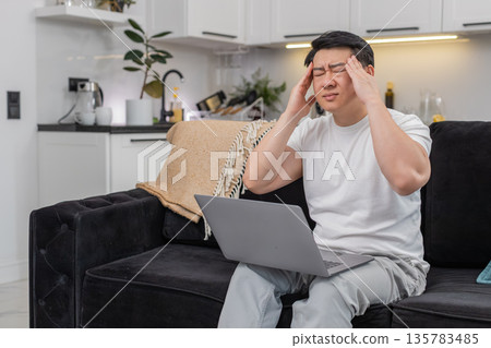 Asian man with laptop feels headache rubs temples closes eyes breathes slowly takes short work break Asian man with laptop feels headache rubs temples closes eyes breathes slowly takes short work break 135783485