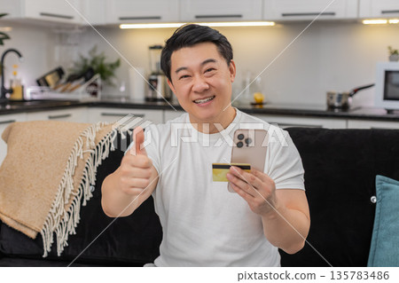 Asian man completes payment with smartphone and credit card smiles at confirmation shows thumbs up Asian man completes payment with smartphone and credit card smiles at confirmation shows thumbs up 135783486