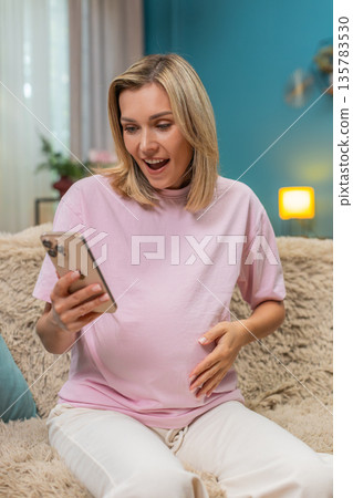 Pregnant woman celebrating baby news texting on smartphone making victory sign smiling super indoors Pregnant woman celebrating baby news texting on smartphone making victory sign smiling super indoors 135783530
